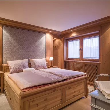 Ein gemütliches Schlafzimmer mit Holzwänden und einem großen Doppelbett. Sanfte Beleuchtung und Fenster mit Vorhängen sorgen für eine einladende Atmosphäre.