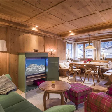 Ein gemütliches Wohnzimmer mit Holzverkleidung und einem großen Esstisch. Es gibt eine grüne Couch, einen runden Tisch und viel natürliches Licht durch die Fenster.