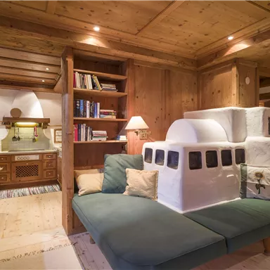 Eine gemütliche Wohnküche mit Holzregalen und warmem Licht. Im Vordergrund befindet sich ein grünes Sofa mit einem auffälligen, skulpturalen Element.