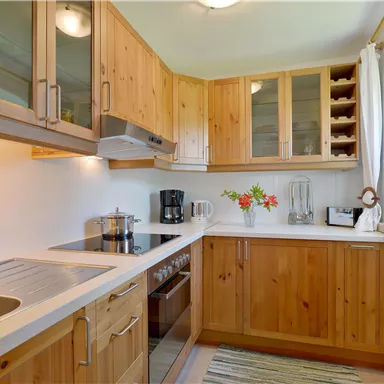 Eine helle Küche mit Holzmöbeln und modernen Geräten. Auf der Arbeitsfläche stehen frische Blumen und verschiedene Küchenutensilien.