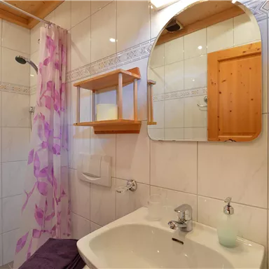 A modern bathroom with a sink and a large mirror. The walls are tiled and a shower is fitted with a curtain partition.