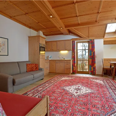 A bright living room with a wooden ceiling and a red carpet. There is a couch, a kitchenette, and a dining area with a window.