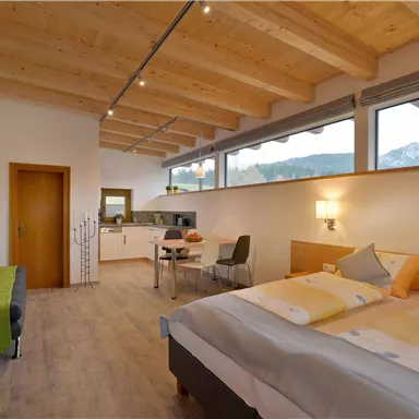 Ein modernes Zimmer mit einem Doppelbett und einem kleinen Essbereich. Große Fenster bieten einen Blick auf die Berge.