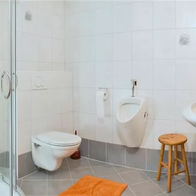 Ein modernes Badezimmer mit Dusche, Toilette und Urinal. Der Boden ist grau gefliest und ein orangefarbener Handtuch liegt auf dem Boden.