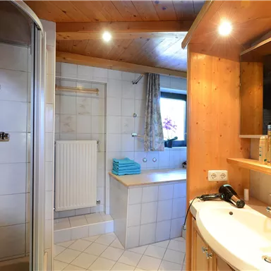 Ein modernes Badezimmer mit einer Dusche und einem Waschbecken. Es gibt helle Fliesen und Holzverkleidung, sowie Handtücher auf dem Regal.