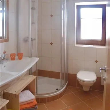 Ein modernes Badezimmer mit einer Dusche, einem Waschbecken und einer Toilette. Die Wände sind hell gefliest, und es gibt einige Handtücher auf einem Regal.