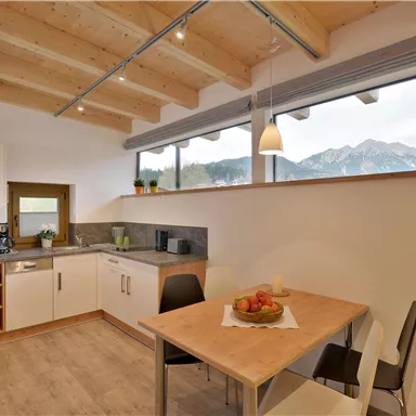 Eine moderne Küche mit Holzdecke und großen Fenstern. Am Tisch stehen frische Früchte und der Blick auf die Berge ist gut sichtbar.