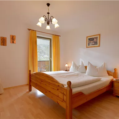 Ein gemütliches Schlafzimmer mit einem Holzbett und zwei Kissen. Das Zimmer hat große Fenster mit gelben Vorhängen und warmem Licht.