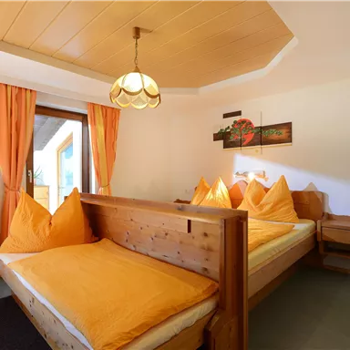 Ein gemütliches Zimmer mit zwei Betten und orangefarbenen Kissen. Die Atmosphäre ist hell und einladend, mit einem Fenster, das Tageslicht hereinlässt.