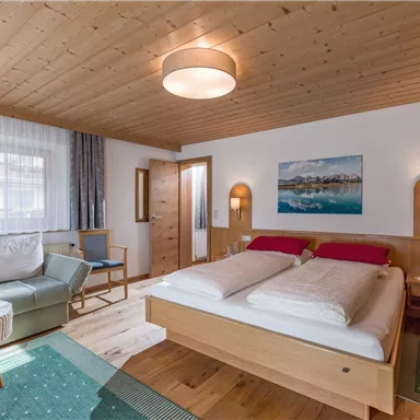 Ein gemütliches Zimmer mit einem Doppelbett, einer Sitzecke und einem holzverkleideten Decke. Große Fenster sorgen für viel Tageslicht und eine angenehme Atmosphäre.