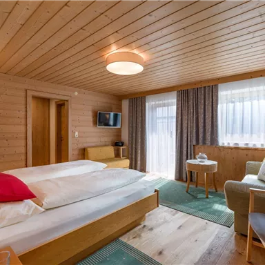 Ein gemütliches Hotelzimmer mit holzverkleideten Wänden und einem großen Fenster. Es ist mit einem Doppelbett, einem Fernseher und einem gemütlichen Sessel eingerichtet.