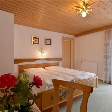 Ein gemütliches Schlafzimmer mit einem Doppelbett und einem Holzdecken. Die Fenster sind mit Vorhängen versehen und es gibt eine Blumendekoration auf dem Tisch.