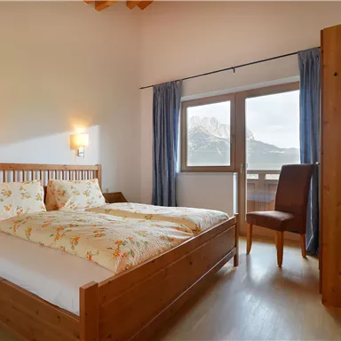 A bright bedroom with a double bed and floral bedding. In front of the window, there is a view of the mountains.