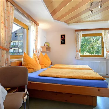 Ein gemütliches Schlafzimmer mit zwei Betten und orangefarbenen Kopfkissen. Der Raum hat große Fenster, durch die viel Tageslicht fällt.