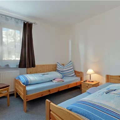 Ein einfaches Schlafzimmer mit zwei Einzelbetten und einem Stuhl. Es gibt ein Fenster mit Vorhängen und eine Nachttischlampe.