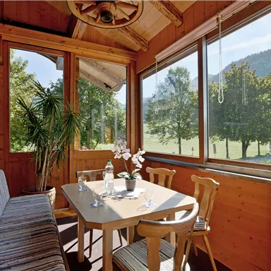 Ein einladender Wintergarten mit Holzverkleidung und stilvollem Tisch. Große Fenster bieten einen Blick auf die grüne Landschaft und Bäume.