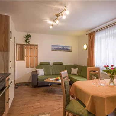 Eine gemütliche Wohnung mit einer Küche, einem Essbereich und einem Wohnzimmer. Die hellen Vorhänge und die Blumen auf dem Tisch verleihen dem Raum eine einladende Atmosphäre.