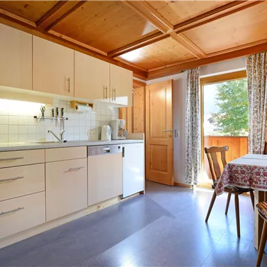 Eine helle Küche mit modernen Möbeln und Holzdecke. Ein kleiner Esstisch steht neben einem Fenster mit Ausblick ins Freie.