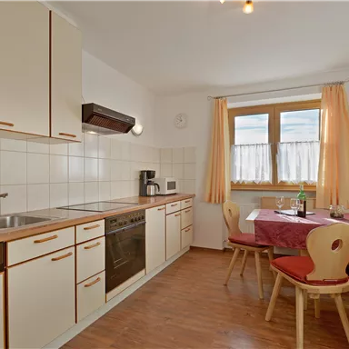 Eine moderne Küche mit weißen Schränken und Holzdetails. Ein Esstisch mit roten Stühlen steht neben einem Fenster mit Vorhängen.