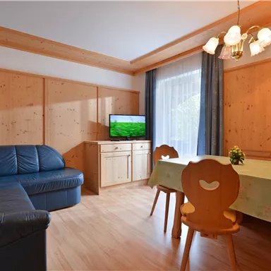 A cozy living room with a blue couch and a table. A television is mounted on the wall, and large windows let in plenty of light.