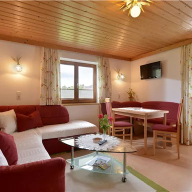 Ein gemütliches Wohnzimmer mit einer roten Couch und Holzbalkendecke. Ein Tisch mit Stühlen und Fenster mit Blick nach draußen sind ebenfalls vorhanden.