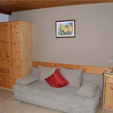 Ein Wohnzimmer mit einem hellen Sofa und einem roten Kissen. Im Raum befinden sich ein Holzschrank, ein Tisch mit einer Lampe und ein Fernseher.