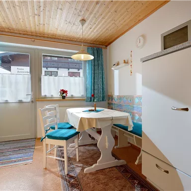 A cozy kitchen with a table and chairs. The room has light-colored walls, a wooden ceiling, and large windows.