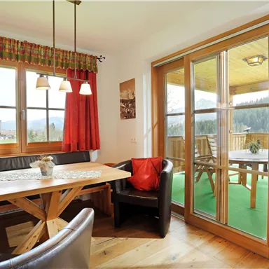 Ein gemütlicher Essbereich mit einem Holztisch und einer dunklen Bank. Große Fenster geben einen Blick auf die Terrasse und die umliegende Natur frei.