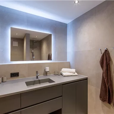 An elegant bathroom with modern amenities and a bright mirror. Towels hang on the wall, and the walls are painted in neutral tones.