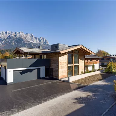 A modern house with a wooden and glass facade in a picturesque mountain landscape. The sky is clear and the surroundings are green and well-kept.
