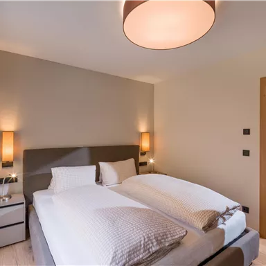 A modern bedroom with a large double bed and soft lighting. The walls are painted in neutral colors, and a wooden closet door is open.