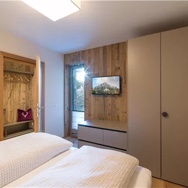 A modern bedroom with two beds and a large window. The walls are partially made of wood, and a television is mounted on the wall.