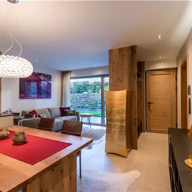 A modern living room with wooden paneling and pleasant lighting. A dining table with a red table runner and a cozy seating area are visible.