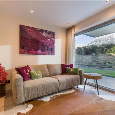 A modern living room with a beige sofa and colorful cushions. Large windows provide a view of the garden and the surrounding nature.