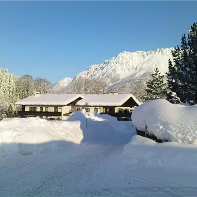 Eine verschneite Landschaft mit einem gemütlichen Haus und schneebedeckten Bäumen. Im Hintergrund sind majestätische Berge unter einem klaren blauen Himmel zu sehen.