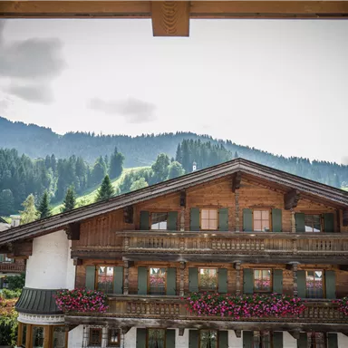 Ein traditionelles Holzhaus mit bunten Blumenbalkonen und Blick auf die Berge. Der Himmel ist bewölkt und die Umgebung ist grün und hügelig.