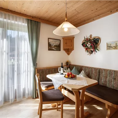 Ein gemütlicher Essbereich mit Holzdekor und einer Sitzecke. Auf dem Tisch liegen Früchte und eine Flasche Wein, während das Licht durch die Fenster strömt.