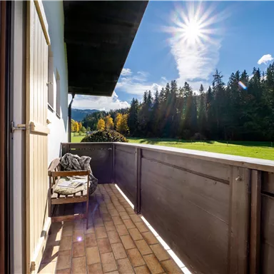 Ein sonniger Balkon mit Holzgeländer und einem Stuhl. Im Hintergrund sind grüne Wiesen und Bäume zu sehen.