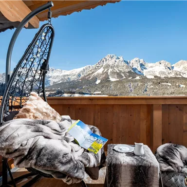 Ein gemütlicher Balkon mit einem schwingenden Stuhl und einer Decke. Im Hintergrund sieht man schneebedeckte Berge und einen strahlend blauen Himmel.