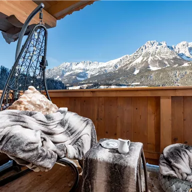 Ein gemütlicher Balkon mit einem schwingenden Stuhl und einer Decke. Im Hintergrund erstrecken sich majestätische Berge unter klarem blauen Himmel.