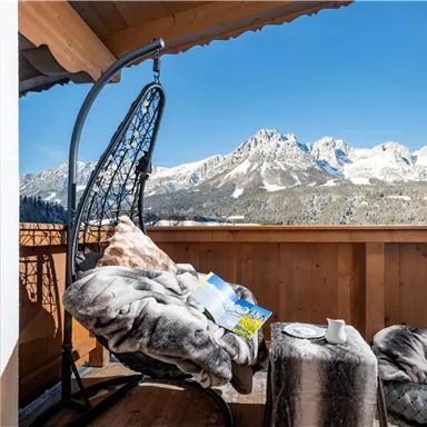Ein gemütlicher Balkon mit einem hängenden Stuhl und einem schönen Ausblick auf schneebedeckte Berge. Auf dem Tisch liegen ein Buch und eine Tasse, ideal zum Entspannen.