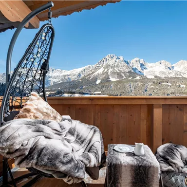 Ein gemütlicher Balkon mit einem schwingenden Stuhl und einer Decke. Im Hintergrund sind schneebedeckte Berge und ein klarer blauer Himmel zu sehen.