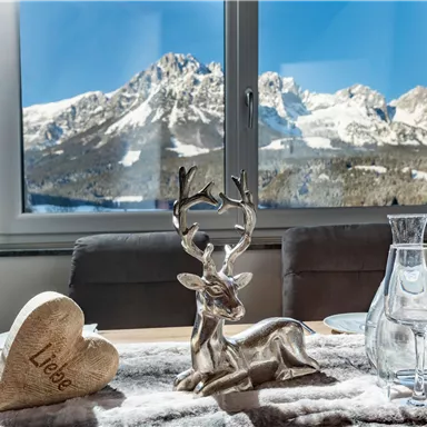 Ein schön gedeckter Tisch mit einem Holzherz, das "Liebe" sagt, und einer kleinen Hirschfigur. Im Hintergrund sind schneebedeckte Berge und ein klarer blauer Himmel zu sehen.