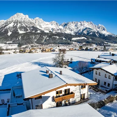 A picturesque winter landscape with snow-covered houses. In the background, majestic mountains rise under a clear blue sky.
