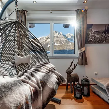 Ein gemütlicher Wohnraum mit einem hängenden Kokonstuhl und Blick auf die Berge. Warmes Licht strömt durch die Fenster und schafft eine einladende Atmosphäre.