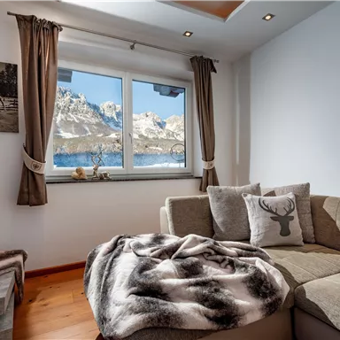 Ein gemütliches Wohnzimmer mit einem grauen Sofa und einem weichen Überwurf. Durch das Fenster ist eine beeindruckende Berglandschaft zu sehen.