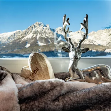 Ein dekorativer Hirsch aus Silber sitzt auf einem weichen, grauen Überwurf. Im Hintergrund sind schneebedeckte Berge und ein blauer Himmel zu sehen.