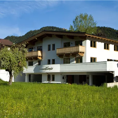 Ein charmantes Gebäude mit Holzbalustraden, umgeben von grünem Gras und Bäumen. Die sonnige Umgebung vermittelt eine einladende Atmosphäre.