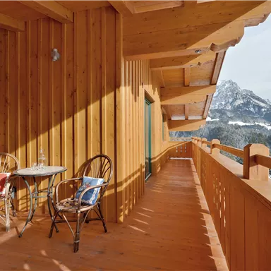 Ein Holzbalcony mit zwei Stühlen und einem Tisch. Im Hintergrund sind schneebedeckte Berge sichtbar.