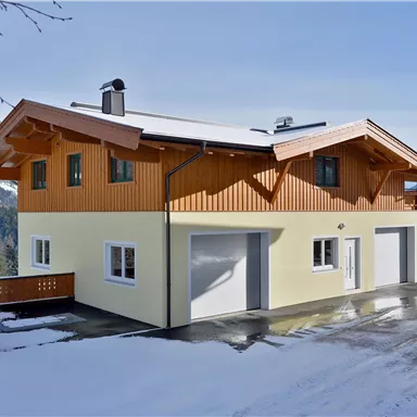 Ein modernes Haus in einer schneebedeckten Landschaft. Im Hintergrund sind Berge und ein klarer Himmel zu sehen.